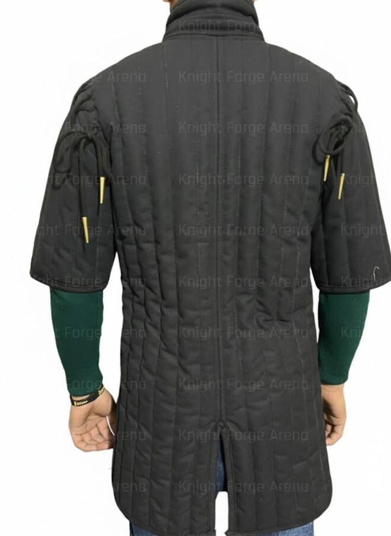 May include: Black padded gambeson with short sleeves and a high collar. The quilted fabric features vertical stitching. The sleeves have decorative gold-colored accents. The back of the garment is visible, showcasing the design and construction.