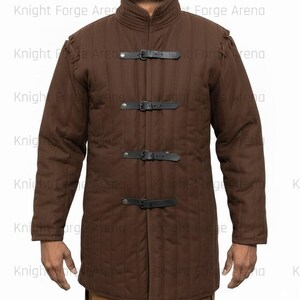 May include: Brown quilted jacket with black leather straps and buckles. The jacket has a high collar and long sleeves. The text "Knight Forge Arena" is visible in the background. This garment is suitable for historical reenactment or costume.