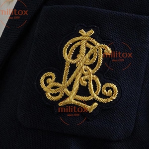 May include: Close-up of navy blue fabric featuring a gold embroidered monogram. The monogram is a stylized "RL" design with detailed gold thread. The fabric has a textured appearance, and the embroidery is raised.