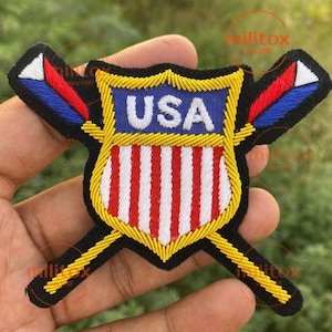 May include: Embroidered patch featuring a shield design with the text "USA" in white on a blue background. The shield has red and white stripes, and is outlined in gold. Two crossed flags are on either side of the shield.
