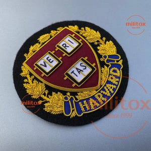 以下が含まれることがあります： ハーバード大学の紋章が刺繍されたパッチ。紋章は、"VE RI TAS"の文字が入った3つの白い箱が付いた、マルーン色の盾です。パッチは金色の刺繍で囲まれ、青色で「HARVARD」の文字が施されています。
