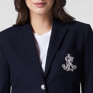 May include: Navy blue blazer with a silver emblem on the pocket and a decorative button. The blazer is worn over a white t-shirt. The blazer's fabric has a subtle texture.
