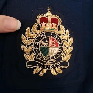 Może przedstawiać: Haftowana naszywka z motywem herbu i napisem "Ralph Lauren" w okrągłym wzorze. Herb zawiera koronę, liście laurowe i tarczę z czerwonymi, białymi, zielonymi i złotymi akcentami. Naszywka jest przyszyta do granatowej tkaniny.