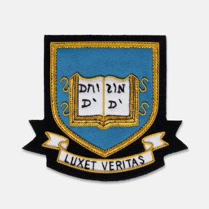 May include: Embroidered patch featuring a shield design with a light blue background and a gold border. The shield contains an open book with text and the words "LUX ET VERITAS" on a white banner below. The patch is black around the edges.