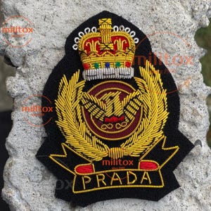 Puede incluir: Un parche bordado negro con una corona dorada, un emblema de águila dorada y la palabra "PRADA" en letras doradas. El parche tiene detalles dorados y rojos. El parche es probablemente un emblema decorativo.