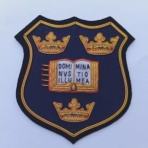 May include: A navy blue embroidered patch in a shield shape, featuring three gold crowns and an open book with the text "DOMI MINA NVS TIO ILLU MEA". The patch has a gold border and black trim.