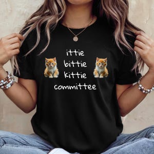 Op de afbeelding: Zwart T-shirt met de witte tekst "ittie bittie kittie committee". Twee oranje tabby kittens staan aan weerszijden van de tekst. Ronde hals.