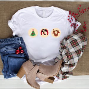 May include: White t-shirt featuring a Christmas tree, snowman, and reindeer design. The shirt is laid out on a brown surface with blue jeans, brown boots, and a plaid scarf.