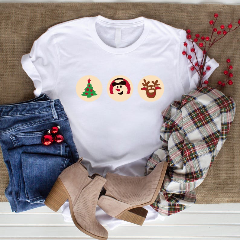 May include: White t-shirt featuring a Christmas tree, snowman, and reindeer design. The shirt is laid out on a brown surface with blue jeans, brown boots, and a plaid scarf.