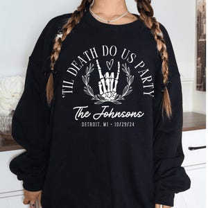 Puede incluir: Sudadera negra con texto blanco que dice "Til Death Do Us Party" con una mano de esqueleto haciendo un signo de rock and roll dentro de una corona. El texto "The Johnsons" est&aacute; debajo de la corona con el texto "Detroit, MI - 10/29/24" debajo.