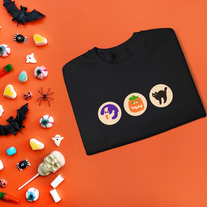 May include: Black t-shirt with three colourful iron-on patches: a ghost, a pumpkin, and a black cat. The patches are arranged horizontally across the front of the shirt.