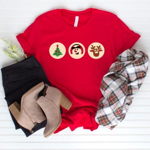 May include: Red t-shirt featuring three Christmas icons: a green Christmas tree, a snowman wearing a black hat, and a reindeer with brown antlers. The icons are displayed in white circles on the red shirt.