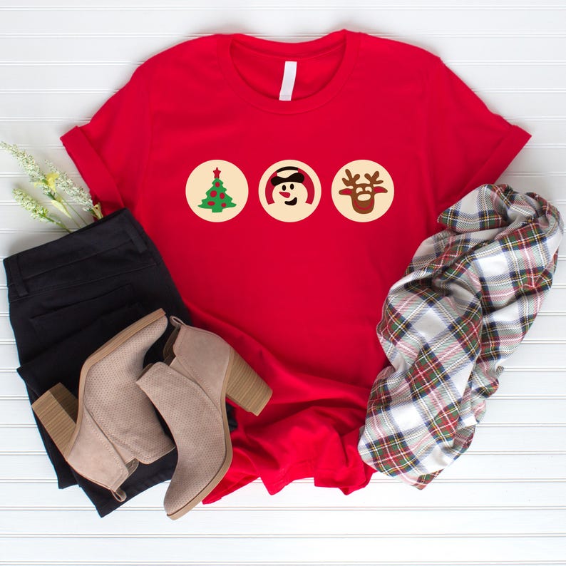 May include: Red t-shirt featuring three Christmas icons: a green Christmas tree, a snowman wearing a black hat, and a reindeer with brown antlers. The icons are displayed in white circles on the red shirt.