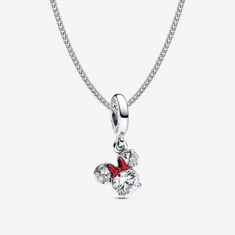 May include: A silver necklace featuring a dangling charm shaped like a mouse head with two clear round stones for ears and a larger clear round stone for the face, accented by a small red enamel bow between the ears on a delicate silver chain.