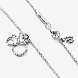 Op de afbeelding: Een zilverkleurige ketting met een Minnie Mouse-silhouet hanger met een enkele heldere steen in de strik. De ketting bevat een karabijnsluiting, een rechthoekig label met de inscriptie PANDORA en een kleine bedel in de vorm van het Disney-logo.
