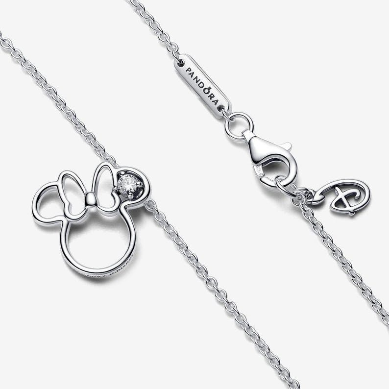 Op de afbeelding: Een zilverkleurige ketting met een Minnie Mouse-silhouet hanger met een enkele heldere steen in de strik. De ketting bevat een karabijnsluiting, een rechthoekig label met de inscriptie PANDORA en een kleine bedel in de vorm van het Disney-logo.