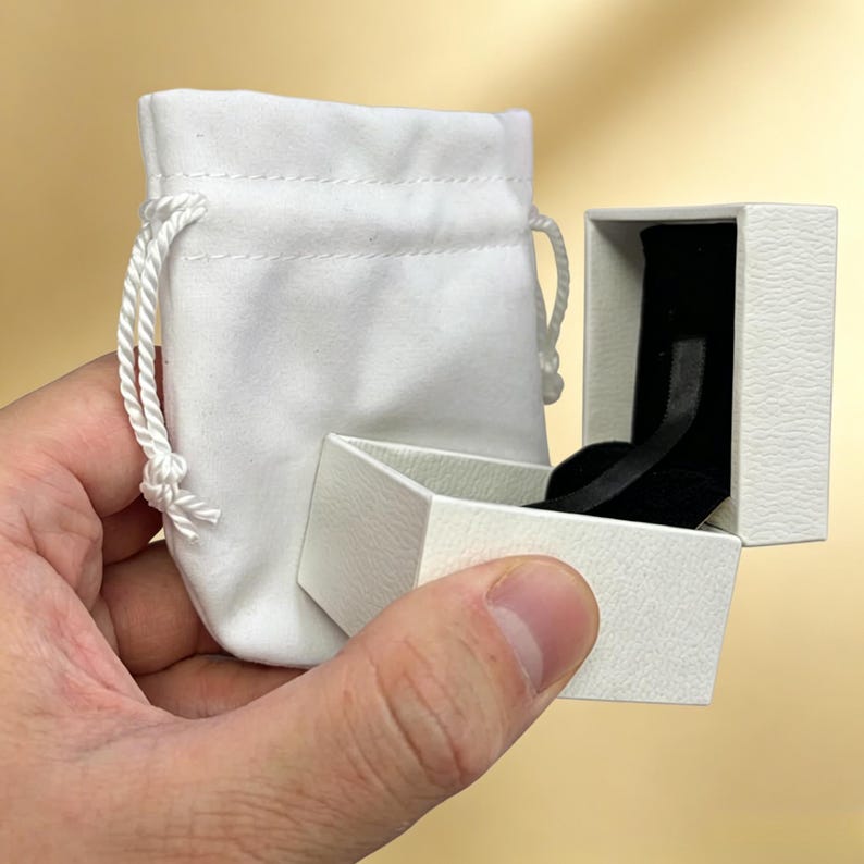 Op de afbeelding: Een wit stoffen zakje met wit trekkoord, gecombineerd met een open wit juwelendoosje met een getextureerde buitenkant en een zwarte fluwelen voering. Het doosje is rechthoekig van vorm en is voorzien van een scharnierend deksel.