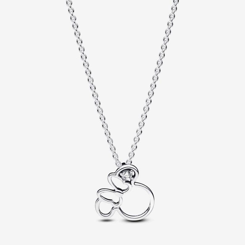 Op de afbeelding: Een zilverkleurige ketting met een hanger in de vorm van een gestileerd muizenhoofdsilhouet met een strik. De hanger bevat een enkele heldere, rondgeslepen edelsteen in het bovenste gedeelte van het ontwerp, hangend aan een fijne ketting.
