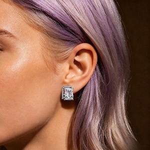 May include: A close-up of a silver stud earring with a large, rectangular, clear gemstone. The earring is surrounded by smaller, clear gemstones. The earring is on a person's ear, with purple hair in the background.