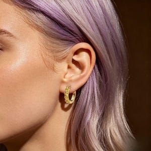 May include: A small gold hoop earring featuring a row of bright yellow, faceted stones set along the outer edge. The hoop has a polished metallic finish and a hinged closure mechanism, securely fastened through the earlobe with a visible clasp.