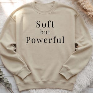Puede incluir: Sudadera beige de cuello redondo con las palabras "Soft but Powerful" impresas en negro. La sudadera tiene mangas largas y cuello redondo. El fondo incluye elementos decorativos.