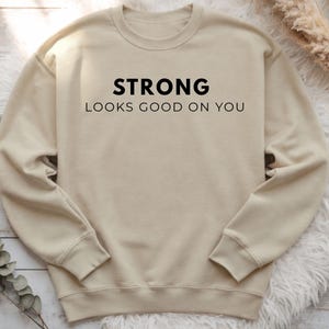 Puede incluir: Una sudadera beige con el texto "STRONG LOOKS GOOD ON YOU" en negro. La sudadera tiene cuello redondo y mangas largas. El fondo incluye una superficie blanca y esponjosa y plantas secas.