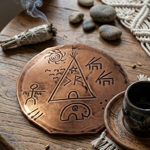 Könnte beinhalten: Eine kupferfarbene, runde dekorative Platte mit eingeätzten Stammeszeichen, darunter ein Tipi, eine Sonne und Vögel. Eine kleine, dunkle Keramiktasse steht auf einer passenden Untertasse. Ein Bündel brennenden Salbeis und kleine Steine sind ebenfalls vorhanden.