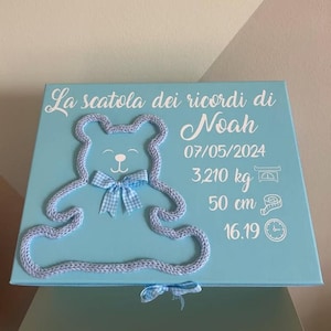 Può includere: Una scatola ricordo azzurra con un contorno di orsetto in maglia e un fiocco a quadretti. La scatola riporta il nome "Noah" e i dettagli della nascita: 07/05/2024, 3.210 kg, 50 cm e 16.19. È presente anche il testo "La scatola dei ricordi di".
