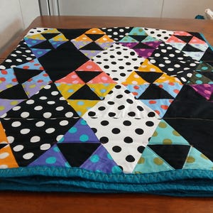 May include: A colorful patchwork quilt with a geometric design. The quilt features triangles and squares in black, white polka dots, and various colors like orange, yellow, purple, and teal. The quilt is trimmed with a teal border.