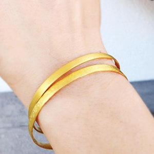 Puede incluir: Dos pulseras de oro en la muñeca de una persona. Las pulseras son finas y tienen una textura martillada.
