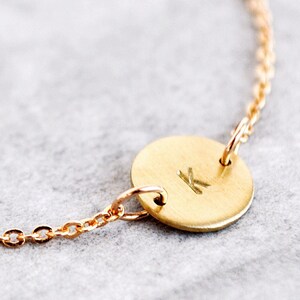 Könnte beinhalten: Ein zartes goldfarbenes Armband mit Kettendesign. Das Armband hat einen kleinen, runden Anhänger mit dem eingravierten Buchstaben "K". Das Armband ist verstellbar und liegt auf einer grauen Oberfläche.