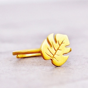 May include: A gold-colored ring with a leaf-shaped design. The leaf is a simple, stylized design with a single vein running down the center.