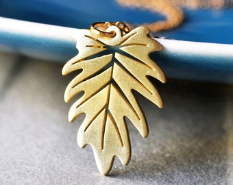 May include: A gold leaf pendant necklace with a delicate, intricate design. The pendant is shaped like a leaf with a detailed vein pattern. The necklace is hanging on a blue plate.