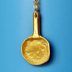 Può includere: Una collana con pendente dorato con un ciondolo a forma di cucchiaio. Il ciondolo ha una superficie strutturata ed è sospeso a una delicata catena d'oro. Lo sfondo è blu brillante.