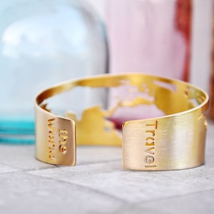 Può includere: Bracciale a polsino color oro con le parole "Travel the World" ritagliate. Il bracciale presenta un design a mappa del mondo. Lo sfondo include bottiglie di vetro sfocate blu e rosa.