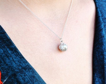 May include: A delicate silver necklace featuring a small, round pendant with a textured surface. The pendant hangs from a fine silver chain. The necklace is displayed against a dark blue floral patterned background.