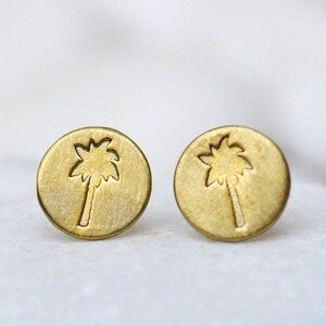 Puede incluir: Dos pendientes de color dorado con un diseño de palmera. Los pendientes están sobre un fondo blanco.