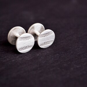May include: A pair of silver cufflinks with a baseball design. The cufflinks are round and have a textured surface.