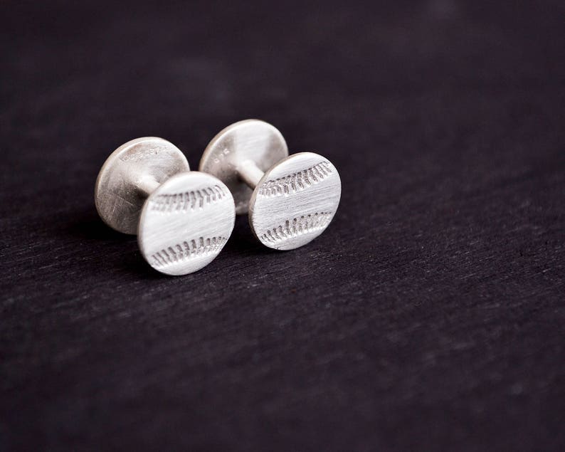 May include: A pair of silver cufflinks with a baseball design. The cufflinks are round and have a textured surface.