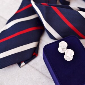 May include: A pair of silver cufflinks with a baseball design, displayed on a blue velvet box. The cufflinks are next to a navy blue and white striped tie with red stripes.