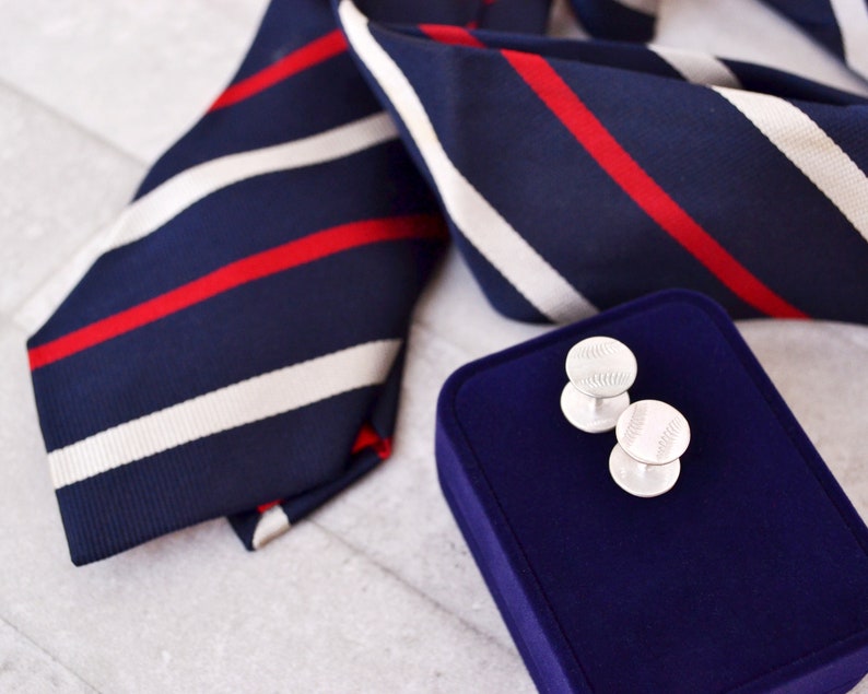 May include: A pair of silver cufflinks with a baseball design, displayed on a blue velvet box. The cufflinks are next to a navy blue and white striped tie with red stripes.