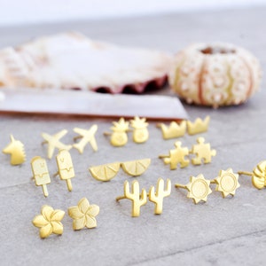 May include: A collection of gold-toned earrings featuring various summer-themed designs, including a pineapple, a crown, a puzzle piece, a lemon slice, a cactus, a sun, a monstera leaf, a plane, an ice cream cone, a flower, and a unicorn.