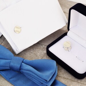 May include: A blue bow tie, a white dress shirt cuff with a gold cufflink, and a black velvet box with a gold cufflink inside.