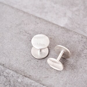May include: A pair of silver cufflinks with a baseball design. The cufflinks are on a gray textured surface.