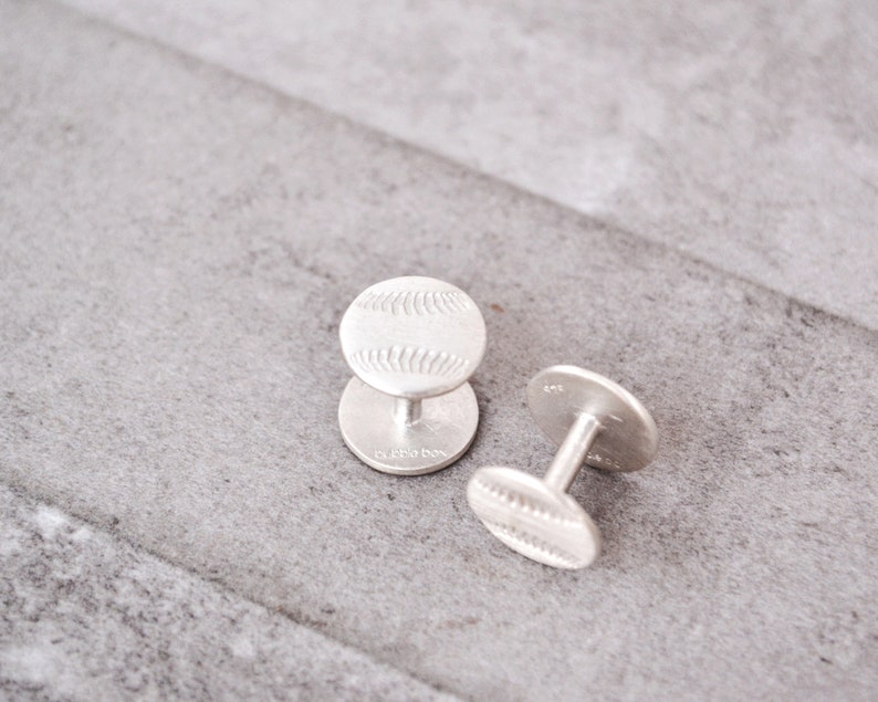 May include: A pair of silver cufflinks with a baseball design. The cufflinks are on a gray textured surface.