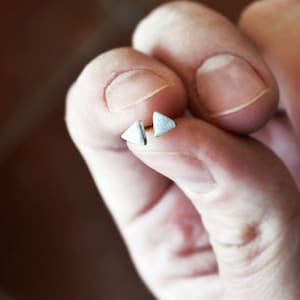 Op de afbeelding: Een paar zilveren driehoekige oorbel studs. De oorbellen zijn klein en delicaat, en worden in de hand van een persoon gehouden.