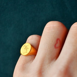 Puede incluir: Un anillo de sello de oro con una cara ovalada que presenta un dibujo lineal de dos figuras bes&aacute;ndose. El anillo se lleva en el dedo de una mano con un coraz&oacute;n rojo dibujado en el dedo.