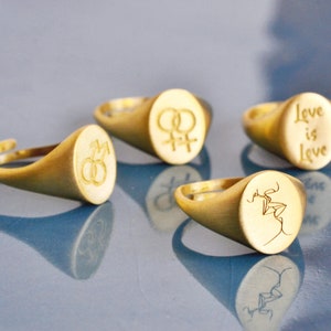 Puede incluir: Cuatro anillos de sello de oro con diferentes grabados. Los anillos presentan los s&iacute;mbolos de masculino y femenino, una pareja bes&aacute;ndose y las palabras "Love is Love".