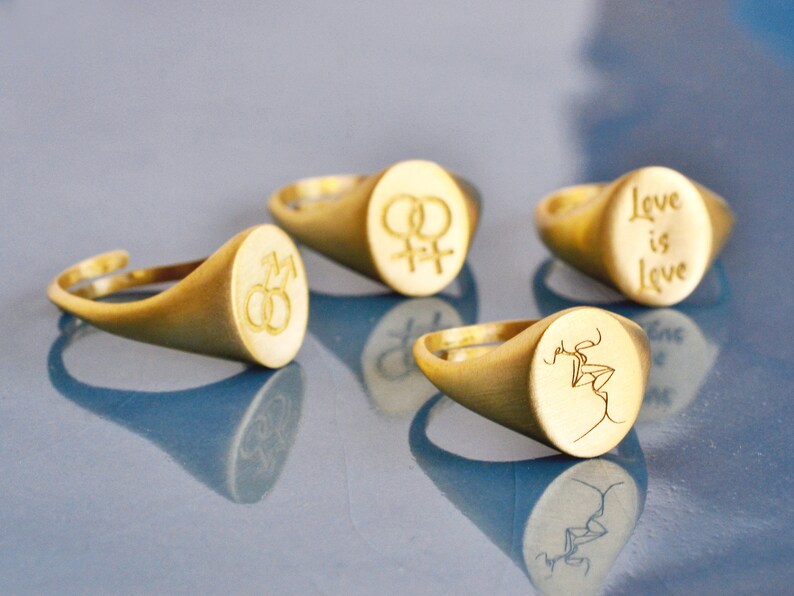 Puede incluir: Cuatro anillos de sello de oro con diferentes grabados. Los anillos presentan los s&iacute;mbolos de masculino y femenino, una pareja bes&aacute;ndose y las palabras "Love is Love".