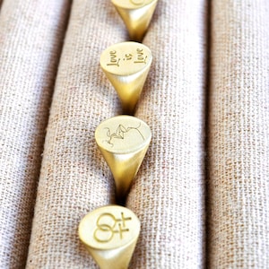 Puede incluir: Cuatro anillos de sello de oro con diferentes grabados. Los anillos se muestran en una fila sobre un fondo de tela beige. Los grabados incluyen las palabras "Love is Love", un par de corazones entrelazados, una imagen estilizada de dos personas bes&aacute;ndose y el s&iacute;mbolo femenino y masculino.
